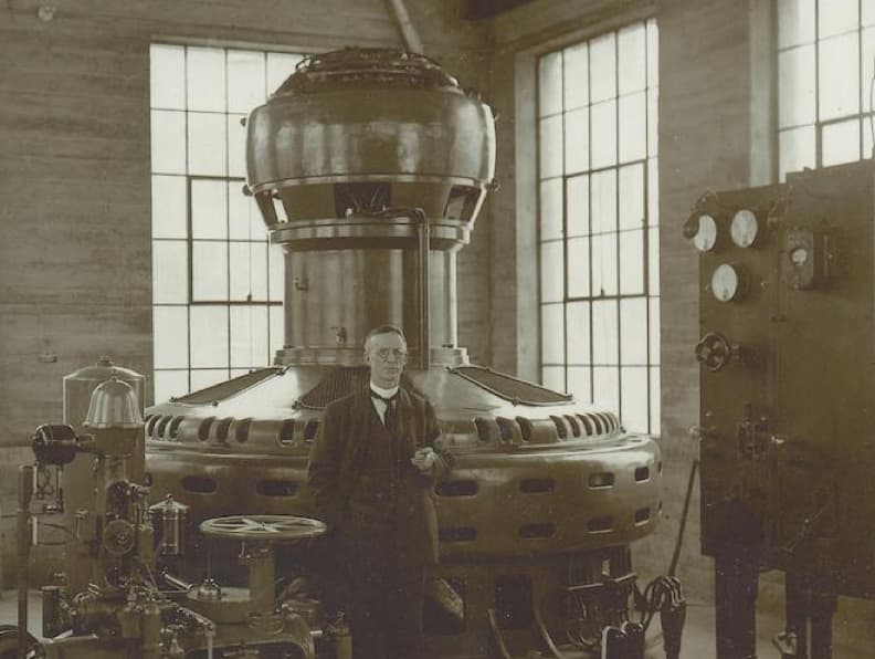 Black and white photograph of a man in a suit standing in a large room with machinery.