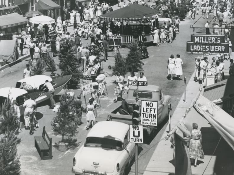 Black and white photograph of Mississaga St. taken from the right corner of West St. and Mississaga St. The street is milling with people and vendors, numerous trees, and the sign for “Miller's Ladies Wear” is clearly visible.