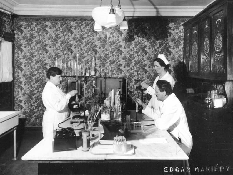 Black and white photograph of a windowless room with floral wallpaper. A lamp hangs from the ceiling, a large cabinet is on the right wall, and a large sink is on the left wall. In the center is a large table with an array of laboratory instruments. Test tubes, beakers, and other less recognizable instruments are handled by two women in nurse's uniforms and a man in a lab coat.