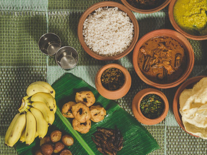 A spread of various traditional dishes on a table with rice, curries, and bananas on a green woven mat. The food is served in earthenware pots.