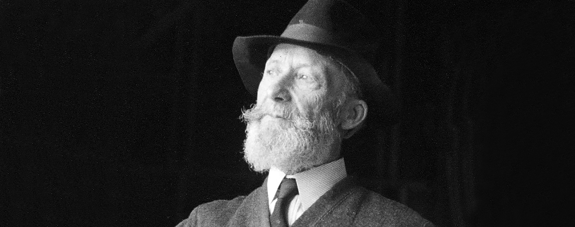 Black and white photograph of Ozias Leduc, sitting, aged 70 to 75. He wears a hat and has a beard. He has his left hand in his trouser pocket.