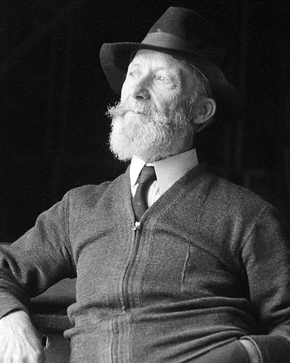 Black and white photograph of Ozias Leduc, sitting, aged 70 to 75. He wears a hat and has a beard. He has his left hand in his trouser pocket.