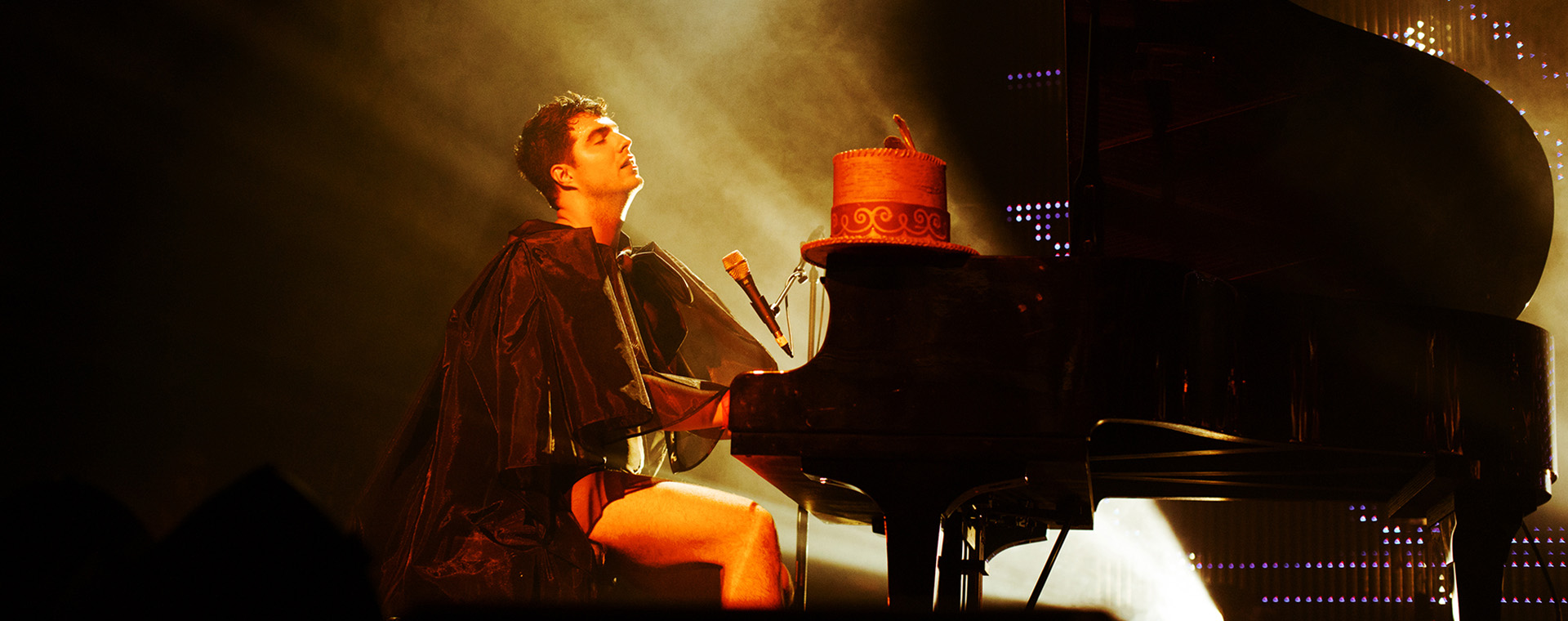 A man is seated at a piano wearing a cape. His tall formal hat rests on the piano. His eyes are closed.