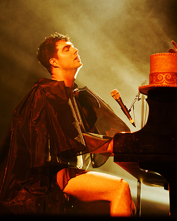 A man is seated at a piano wearing a cape. His tall formal hat rests on the piano. His eyes are closed.