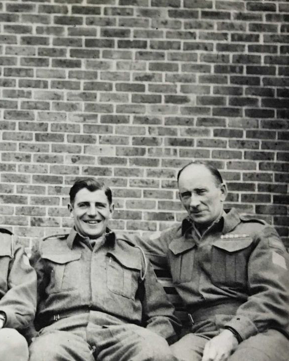 Black and white photograph. Archie, at right, sits on a bench with a much younger man. Both are in uniform and are smiling.