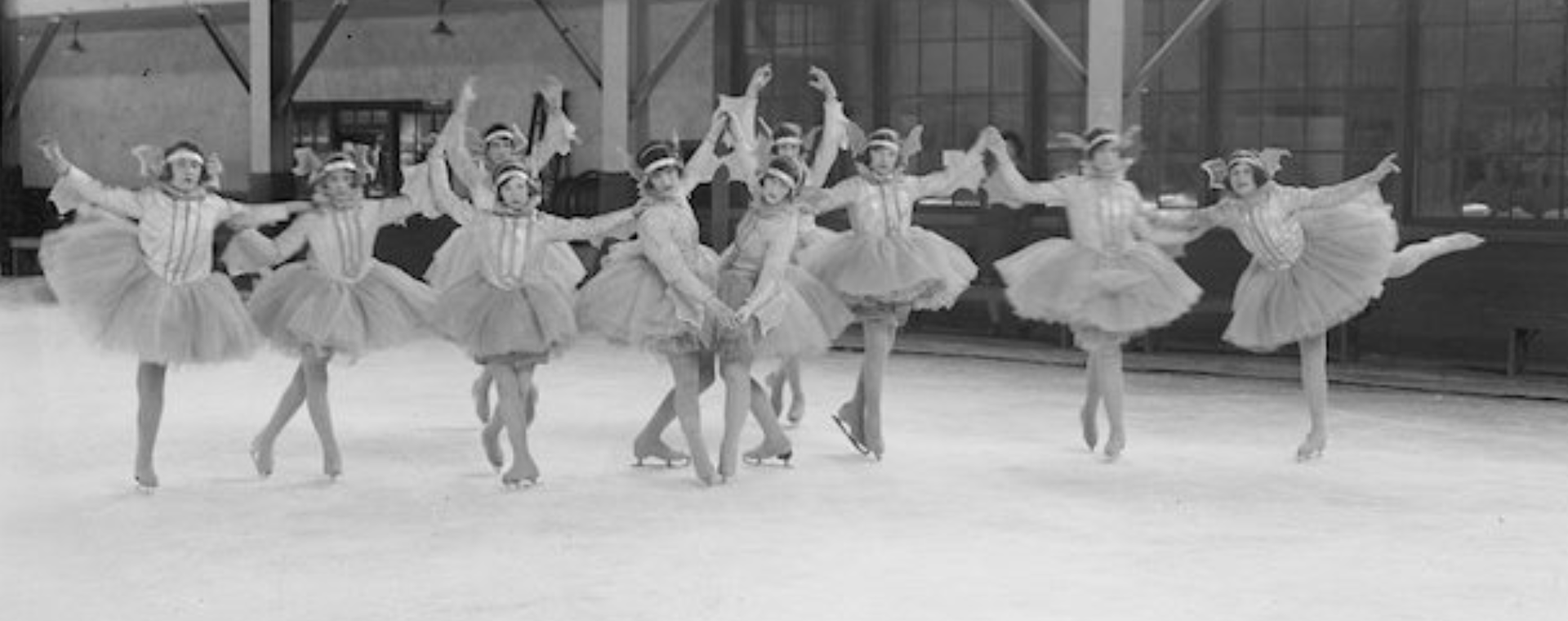 A black and white photograph of ten tutu-costumed skaters in various poses. The group is in an indoor ice rink.