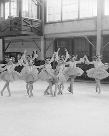 A black and white photograph of ten tutu-costumed skaters in various poses. The group is in an indoor ice rink.