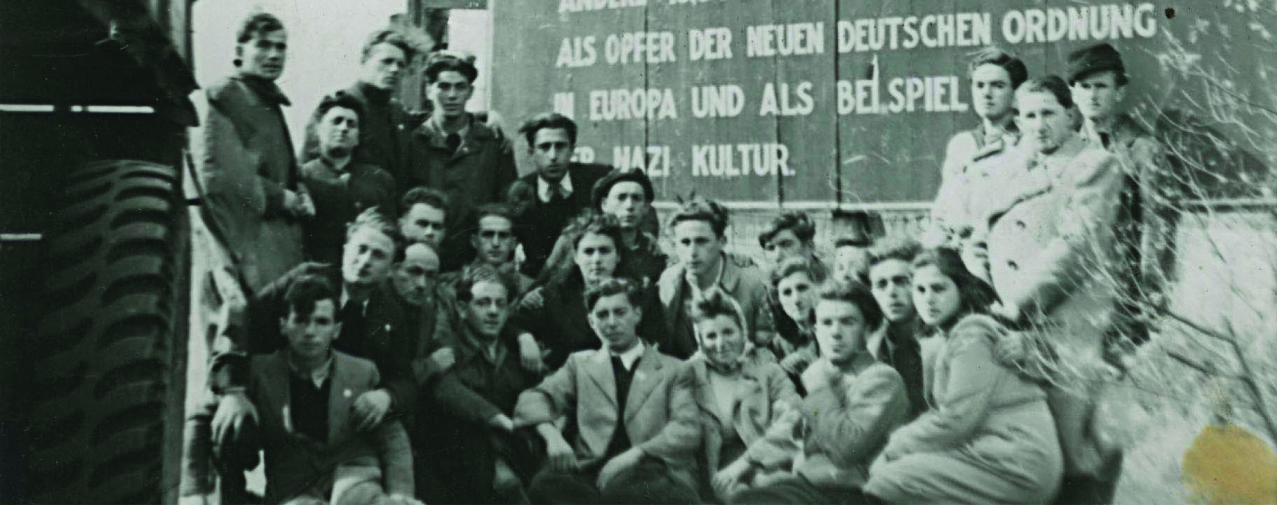 Black-and-white photo: A group of men and women posing in front of sign written in German marking the entrance to Bergen-Belsen Displaced Persons Camp.