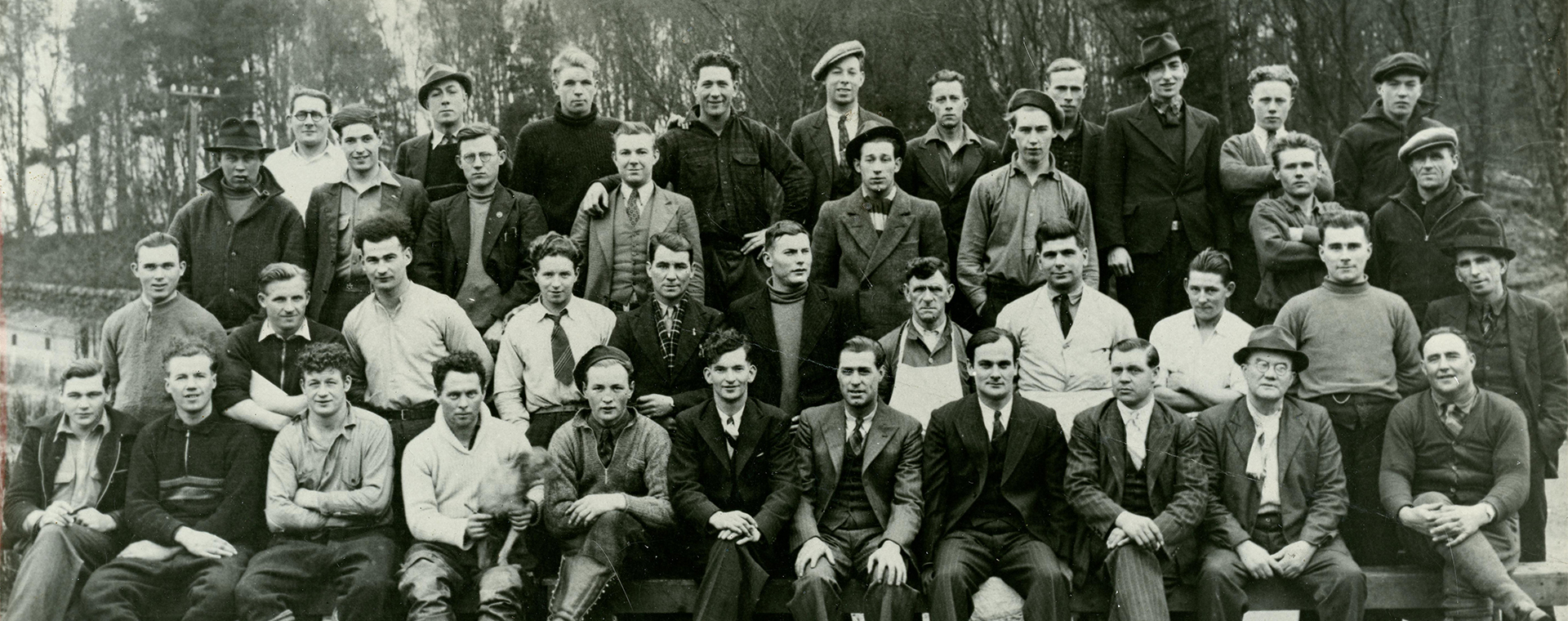 A black and white photograph of 40 men dressed casually or in suits. They sit or stand in 4 rows, posed for an outdoor photo. Many men smile and look at the camera. A row of trees is in the distance behind them.